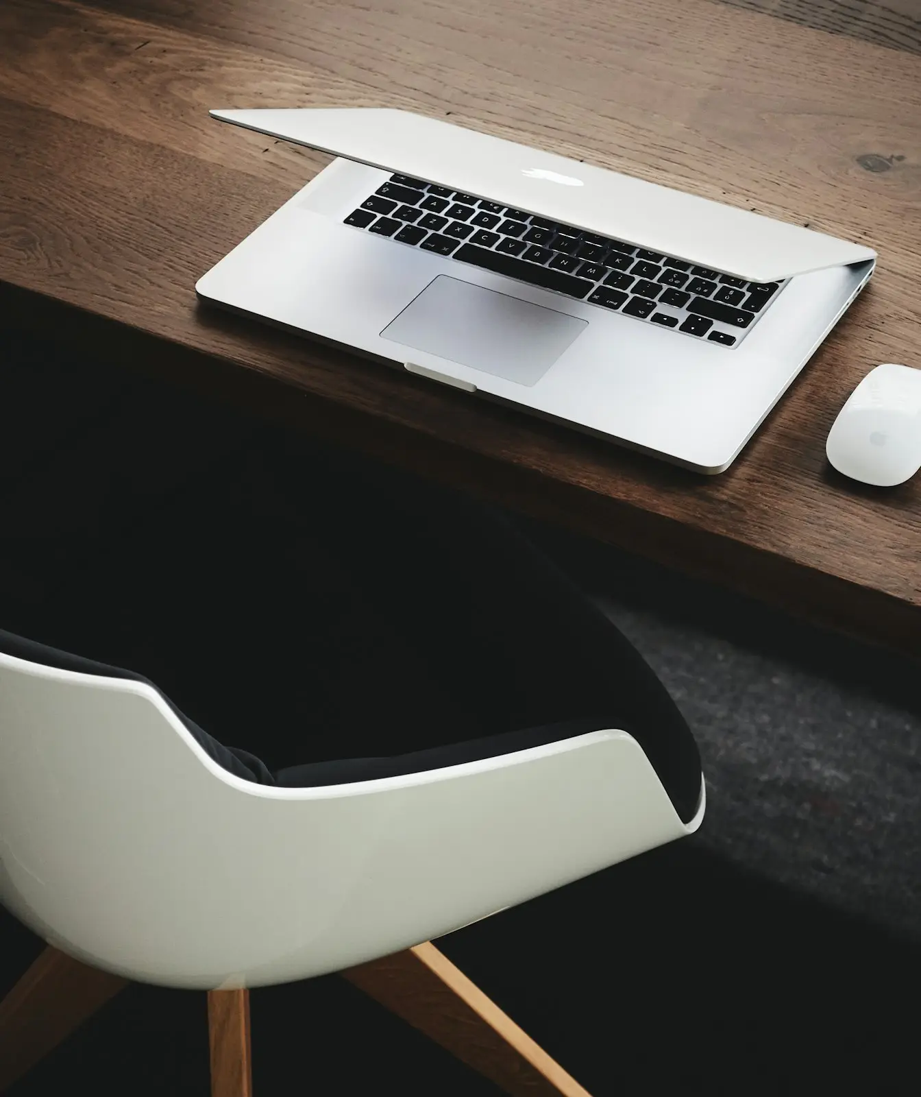 A laptop on a dark wooden desk beside an empty white chair
